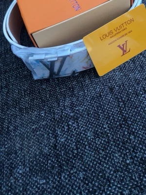 H&M bälte  - Säljer ett snyggt H&M bälte med klassiskt H&M i silver. Bältet har ett färgstarkt mönster med inslag av vitt och andra färger, och levereras med originalask och certifikat. Perfekt accessoar för att lyfta din outfit.