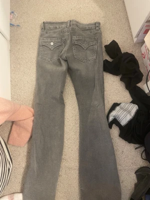 Grå bootcut jeans från Gina trikot - Säljer ett par grå jeans från Gina trickot med bootcut passform. Jeansen har snygga detaljerade bakfickor med dekorativ söm och metallknapp med logga. Klassisk femficksmodell i mjukt denimtyg, perfekta till en avslappnad stil.