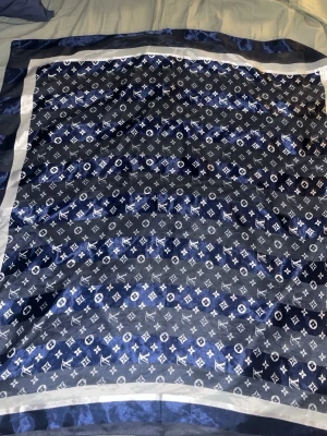  Louis Vuitton scarf  - Snygg blå sjal från Louis Vuitton med klassiskt monogrammönster i vitt och mörkblått. Sjalens material är glansigt och känns lyxigt, och den har en bred ljus kant runt om. Perfekt accessoar för att lyfta din outfit.