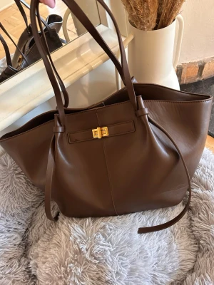 Brun shoppingväska i läderlook - Stilren brun shoppingväska i läderimitation med långa axelband och guldfärgat vridlås framtill. Väskan har en rymlig design och mjuka former, perfekt för att bära allt du behöver. Snygg detalj med knytband på sidorna och minimalistisk look. (Pris går att diskutera)
