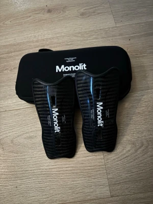 Monolit benskydd  - Säljer ett par svarta benskydd från Monolit i kolfiber med snyggt mönster och vit logga. De har en ergonomisk form och kommer med ett svart fodral med dragkedja. Perfekta för dig som vill ha lätta och tåliga skydd till fotboll. Lite sönder på båda som på bilderna men annars är dom hela! 