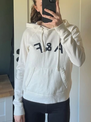 Vit hoodie från Abercrombie & Fitch - Snygg vit hoodie från Abercrombie & Fitch med svart A&F-tryck på bröstet. Hoodien har huva med dragsko och en stor magficka. Perfekt för en avslappnad och trendig look. Mjuk bomullskänsla och klassisk passform.