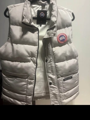 Väst herr och dam - Beige dunväst från Canada Goose med logga på bröst köptes ny för 2 år sedan men endast används fåtal gånger då jag gått upp vikt behöver större stl. Men denna passar S och M. Passar kvinnor som har stl M oxå…