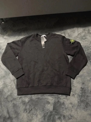 Svart sweatshirt från Stone Island - Säljer en svart sweatshirt från Stone Island med klassisk rund hals och ribbade muddar. Tröjan har den ikoniska Stone Island-loggan på vänster ärm och är tillverkad i mjukt material för en skön passform.