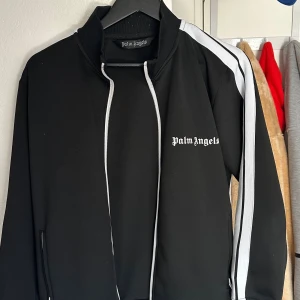 Palm Angels svart track jacket - Säljer en svart track jacket från Palm Angels med vita ränder längs ärmarna och vit logga på bröstet. Jackan har dragkedja framtill, hög krage och ribbade muddar. Perfekt för en streetwear-look. Inte säker på om tröjan är äkta därav priset.