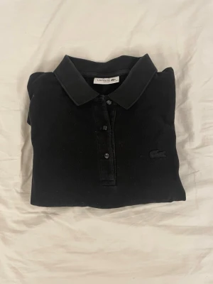 Svart pikétröja från Lacoste - Klassisk svart pikétröja från Lacoste med krage och knappar framtill. Tröjan har den ikoniska krokodilloggan ton-i-ton på bröstet och är gjord i bomull för en bekväm känsla. Perfekt till jeans eller shorts för en clean look.