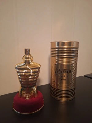 Jean Paul Gaultier Le Male Elixir Herr Parfym - 125 ml  - Exklusiv parfymflaska i guld från Jean Paul Gaultier, Le Male Elixir. Flaskan har en unik kroppsliknande form och står på en röd sammetsbas. Medföljer en metallisk cylindrisk förpackning med märkets logotyp. Perfekt för dig som vill ha något lyxigt och iögonfallande.           ⚡️ Snabba svar på meddelanden 💬 Tveka inte att skicka ett meddelande om du har frågor. 📦 Snabb frakt – skickar samma eller nästa dag efter beställning.  🏷Lägg flera varor i bundle för upp till 20% rabatt.  