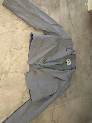 Ljusblå kort kavaj från ZARA - Trendig ljusblå kavaj från ZARA i kortare modell med en knapp framtill. Kavajen har klassisk krage, långa ärmar och är fodrad. Perfekt för att lyfta din outfit med en fräsch färg och stilren look.