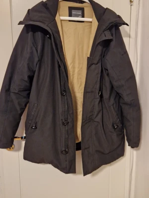 Svart vinter jacka från Race Marines - Snygg svart parkasjacka från Jack & Jones med dragkedja, knappar och justerbar midja. Jackan har flera fickor, huva och beige innerfoder. Perfekt för kalla dagar och har en klassisk, rak passform.   Har haft mindre hål som åtgärdats. Varm jacka! Pris kan diskuteras