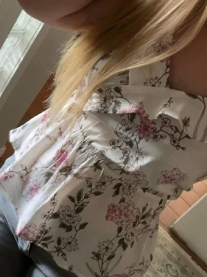 Vit blommig sommarklänning med volang - Söt vit topp med rosa och gröna blommor, omlott framtill och volangdetalj vid urringningen. Toppen har smala axelband och är perfekt för varma dagar. Tillverkad i ett lätt och luftigt material.