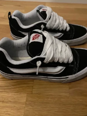 Vans knu skool - Klassiska Vans knu Skool sneakers i svart mocka och canvas med vita detaljer och den ikoniska sidoremsan. Skorna har tjocka vita snören, vit sula och röd Vans-logga på plösen. Perfekta för en streetwear-look. 