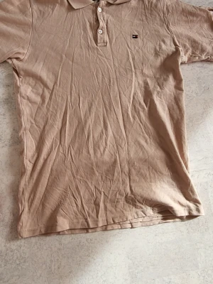 Beige pikétröja från Tommy Hilfiger - Snygg beige pikétröja från Tommy Hilfiger med klassisk krage och knappar framtill. Tröjan har korta ärmar och en liten logga på bröstet. Tillverkad i mjuk bomull som känns skön mot huden. Perfekt för en clean och enkel stil.