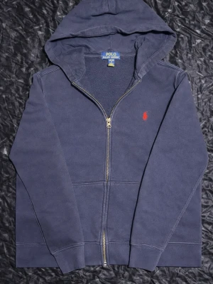 Marinblå hoodie från Polo Ralph Lauren - Snygg marinblå hoodie med dragkedja från Polo Ralph Lauren. Klassisk design med röd broderad logga på bröstet, huva och kängurufickor. Tillverkad i mjuk bomullsblandning som är skön att ha på sig. Perfekt för en avslappnad och stilren look.
