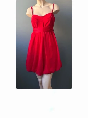 Röd kort klänning med knytband - Snygg röd klänning med tunna axelband och ett brett knytband i midjan. Klänningen har en luftig kjol med lätt volang och är tillverkad i ett tunt, flygigt material som ger en elegant känsla. Perfekt för dig som vill sticka ut med färg. Storlek L men sitter som S. 