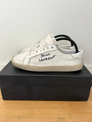 Vita Saint Laurent sneakers med brodyr - Snygga vita sneakers från Saint Laurent med handskriven logga på sidan och broderad text på plösen. Skorna har lågt skaft, klassisk snörning och är tillverkade i canvas med gummisula. Perfekta för en stilren och trendig look. Skicket är hyfsat bra. Pris kan diskuteras 