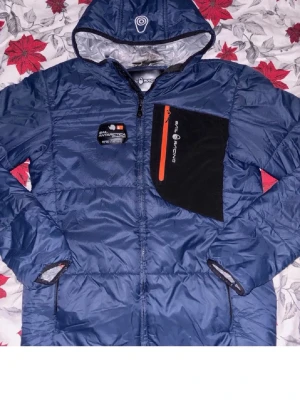 Blå vindjacka från snail raicing - Snygg blå vindjacka med huva från Antarctic. Jackan har en svart bröstficka med orange dragkedja och logga, samt två sidofickor. Materialet är glansigt och lätt, perfekt för blåsiga dagar. Jackan har även detaljer i svart och grått vid ärmslut och huva.