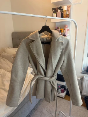 Beige kappa med knytskärp från Vero Moda - Stilren beige kappa från Vero Moda med breda slag och knytskärp i midjan. Kappan har en rak passform och är tillverkad i ett mjukt material som ger en lyxig känsla. Perfekt för dig som vill ha en classy och trendig look.