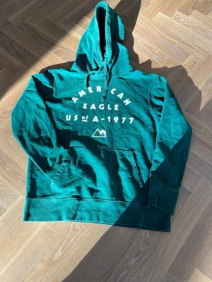  hoodie  American Eagle - Snygg turkos hoodie från American Eagle med vit tryckt text och grafisk detalj framtill. Klassisk känguruficka, dragsko i huvan och mjukt material. Perfekt för en avslappnad och cool stil.