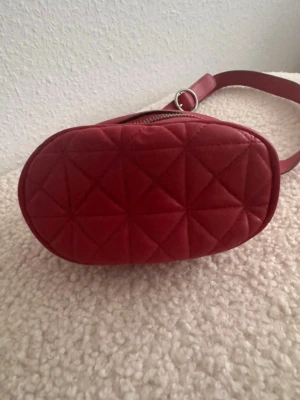 Röd quiltad midjeväska med justerbart band - Snygg röd midjeväska i quiltat mönster med oval form och justerbart axelband. Väskan har dragkedja upptill och är tillverkad i ett mjukt material som ger en trendig look. Perfekt för att bära dina viktigaste grejer med stil. Använd Barsebäck gång.