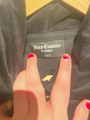 Svart velour hoodie från Juicy Couture - Säljer en svart velour hoodie från Juicy Couture i storlek XXS. Hoodien har dragkedja framtill, två fickor och en broderad JC-logga på bröstet. Klassisk huva och ribbade muddar vid ärmslut och nederkant. Perfekt för en chill och trendig look.