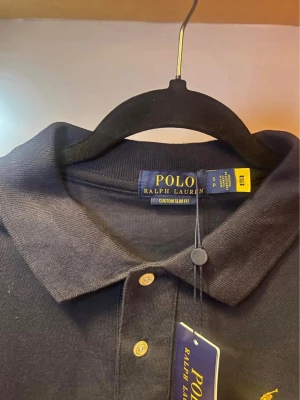 Svart piké från Polo Ralph Lauren - Klassisk svart pikétröja från Polo Ralph Lauren med gul broderad logga på bröstet. Tröjan har korta ärmar, krage och knappar framtill. Modellen är custom slim fit och tillverkad i mjuk bomull som känns skön mot huden.