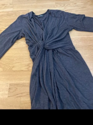 Mörkgrå omlottklänning Club L - Lång mörkgrå omlottklänning från Club L med v-ringning och knyt i midjan. Klänningen har långa ärmar och ett mjukt, stretchigt tyg som ger en snygg siluett. Perfekt för dig som gillar stilrena och bekväma plagg.