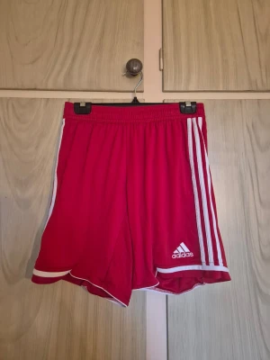 Röda Adidas shorts med vita ränder - Säljer ett par röda Adidas shorts med klassiska vita ränder längs sidorna och logga på framsidan. De har elastisk midja och är tillverkade i lätt polyester, perfekt för sport och träning. Modellen är loose fit och har ClimaCool-teknologi för extra komfort.  Alla kläder rollas innan dom skickas hem då det finns katt i hemmet🐈 Jag stryker och tvättar INTE innan jag skickar det får du stå för själv så det blir som du vill ha det🤗