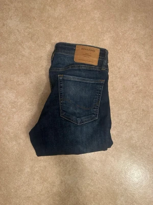 Mörkblå jeans från Jack & Jones - Snygga mörkblå jeans från Jack & Jones 29/30 med klassisk femficksdesign och läderpatch bak i midjan. Jeansen har en normal passform och är tillverkade i bomull med en skön känsla. Perfekta för en stilren och avslappnad look.