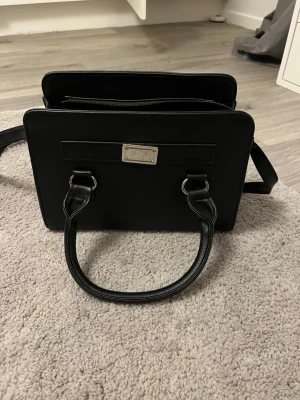 Svart fyrkantig handväska med axelrem - Stilren svart handväska i fyrkantig modell med både handtag och justerbar axelrem. Väskan har en metallbricka framtill och dragkedja upptill. Tillverkad i slätt konstläder och passar perfekt för dig som gillar minimalistisk design. Finns endast ett fack.