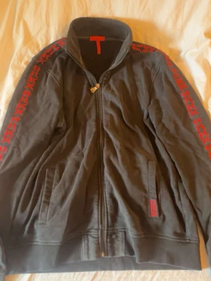 Svart zip-hoodie Hugo boss - Svart hoodie med dragkedja från Hugo boss  dekorerad med röda mönster längs ärmarna och röd logotyp vid fickan. Regular fit i bomull, två fickor framtill och hög krage. Perfekt för en chill och stilren look.