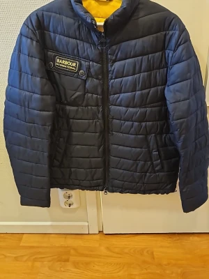 Mörkblå pufferjacka från Barbour - Snygg mörkblå pufferjacka från Barbour International med gul insida. Jackan har quiltad design, dragkedja framtill, bröstficka med Barbour-logga och två sidofickor. Perfekt för kalla dagar och ger en sportig look.