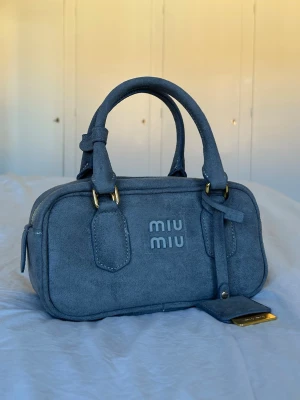 Miu Miu väska - Snygg gråblå handväska från Miu Miu i mjuk mocka med två handtag och guldfärgade metalldetaljer. Väskan har en rektangulär form och logotypen är broderad framtill. Perfekt accessoar för att lyfta din outfit. Nästan oanvänd och i helt nytt skick! Medföljer ett band. 