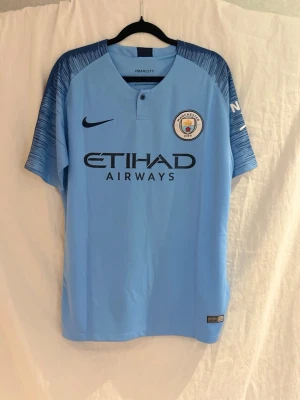 Manchester City matchtröja Sané 19 - Ljusblå Manchester City matchtröja från Nike med Sané och nummer 19 på ryggen. Tröjan har klubbmärke på bröstet, sponsorlogga och mörkblå detaljer på axlarna. Perfekt för fotbollsfans som vill visa sitt stöd på läktaren eller planen.