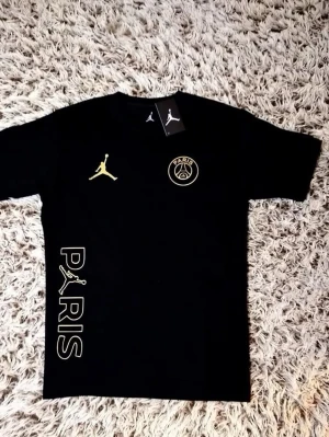 Svart PSG x Jordan t-shirt - Snygg svart t-shirt från Jordan i samarbete med PSG. T-shirten har korta ärmar, guldfärgade loggor på bröstet och texten PARIS med Jumpman-loggan längs sidan. Perfekt för dig som gillar streetwear och sportig stil.