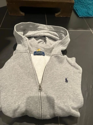 Grå hoodie från Polo Ralph Lauren - Snygg grå hoodie från Polo Ralph Lauren med dragkedja framtill och klassisk huva. Tröjan har det ikoniska lilla broderade logot på bröstet i mörkblått. Tillverkad i mjukt bomullsmaterial som känns skönt mot huden. Perfekt för en avslappnad och stilren look.