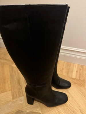 Svarta knähöga stövlar från Diko - Snygga svarta knähöga stövlar från Diko i slätt läder. Modellen har en bred klack och rundad tå, vilket ger en klassisk och stilren look. Perfekta till höstens och vinterns outfits. Dragkedja på insidan för enkel på- och avtagning.