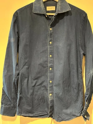 Blå denim skjorta från John Henric - Säljer en klassisk blå skjorta från John Henric med lång ärm och knappar framtill. Skjortan är tillverkad i mjuk bomull och har en enkel, stilren design med normal passform. Perfekt till jeans eller chinos för en clean look.