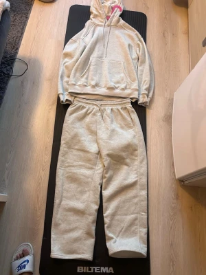 Grått mjukisset med hoodie och byxor - Mysigt loungewear-set i ljusgrått med en oversized hoodie och matchande byxor. Hoodien har stor ficka framtill och dragsko i huvan, medan byxorna har resår i midjan för extra komfort. Perfekt för chill dagar hemma.