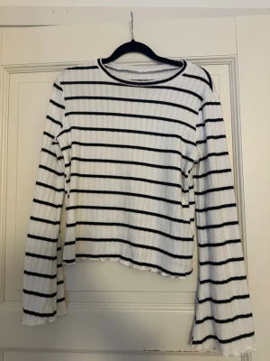 Vit och svart randig ribbad topp - Vit långärmad topp med svarta horisontella ränder och ribbad struktur. Modellen har en något croppad passform och lätt vågiga kanter vid ärmslut och nederkant. Perfekt för en avslappnad och trendig look.