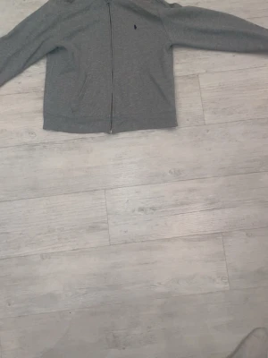 Grå zip hoodie från Polo Ralph Lauren - Snygg grå hoodie med dragkedja från Polo Ralph Lauren. Klassisk design med två fickor fram, liten mörkblå logga på bröstet och huva. Perfekt till chill vardagsstil. Mjuk bomullsblandning som känns skön mot huden.