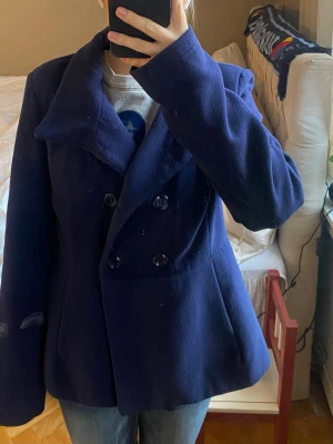 Mörkblå kort kappa med dubbelknäppning - Stilren mörkblå kappa med bred krage och dubbelknäppning framtill. Jackan har två rader med stora svarta knappar och ett dekorativt bälte med knappar i ryggen. Tillverkad i ett tjockare ull-liknande material som håller värmen. Perfekt för kyliga dagar.