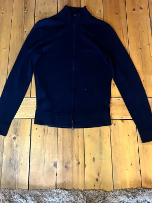 Mörkblå  helzip tröja - Säljer en stilren mörkblå ziptröja med krage och ärmar. Tröjan har hel dragkedja framtill och ribbade muddar vid ärmslut och nederkant. Perfekt att slänga på sig över en t-shirt eller en skjorta  för en clean look. 100% merinoull, märke these glory days. Köpt från volt.