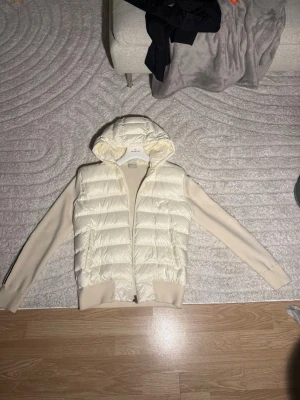 Vit cardigan från Moncler - Snygg vit dunväst från Moncler med stickade ärmar och huva. Västen har dragkedja framtill, ribbade muddar och Monclers ikoniska logga samt randig detalj på ärmen. Perfekt för lager på lager och streetstyle.