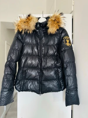 Svart pufferjacka med päls - Snygg svart pufferjacka från italienska Eleven Elfs med stor huva och fluffig päls fylld med dun. Ficka för liftkort på vänster ärm om man vill ha den i backen. Perfekt för kalla dagar och riktigt trendig look.