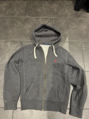 Grå zip hoodie från Polo Ralph Lauren - Mörkgrå hoodie med dragkedja från Polo Ralph Lauren. Klassisk huva med vita snören och röd broderad logga på bröstet. Två fickor framtill och ribbade muddar. Tillverkad i mjuk bomullsblandning för skön känsla.