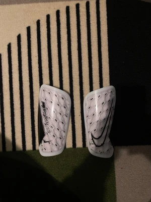 Nike Mercurial benskydd vita - Säljer ett par vita Nike Mercurial benskydd med svart logga och text. De har en lätt, böjd form och mönstrad yta för extra komfort och skydd. Perfekta för fotbollsträning eller match. Snygg och sportig design som passar till all sportutrustning.