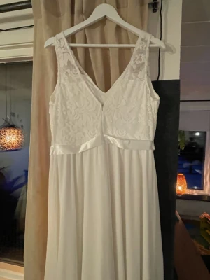 Vit lång spetsklänning med v-ringning - Oanvänd brudklänning från  Strl xl men mer M/L. 150kr, Elegant vit långklänning med v-ringning och breda axelband. Överdelen har vacker spets med blommönster och ett satinband markerar midjan. Kjolen är i ett mjukt, fallande tyg som ger en romantisk look. Perfekt för speciella tillfällen.