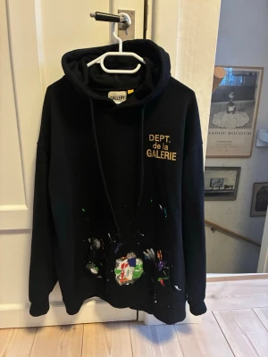 Svart hoodie DEPT. de la GALERIE Gallery Dept. - Svart hoodie från DEPT. de la GALERIE med tryck i gult på bröstet och färgstänk över fickan. Klassisk passform med huva och dragsko. Perfekt för dig som gillar streetwear och unika detaljer.