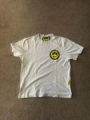 Vit Barrow t-shirt med smileytryck - Säljer en vit t-shirt från Barrow med ett gult smileytryck på bröstet och stor svart BARROW-logga på ryggen. T-shirten har rund hals och korta ärmar. Tillverkad i mjuk bomull och har en avslappnad passform. Perfekt för dig som gillar streetwear och coola prints.