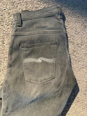 Nudie Jeans - Säljer ett par grå jeans från Nudie Jeans med klassisk femficksmodell och vita vågstickningar på bakfickorna. Jeansen har normal midja, raka ben och är tillverkade i mjuk denim. Perfekta för en avslappnad och stilren look. Jeansen har lite wear and tear på fickorna men utifrån det är den i perfekt skick. W31 L34